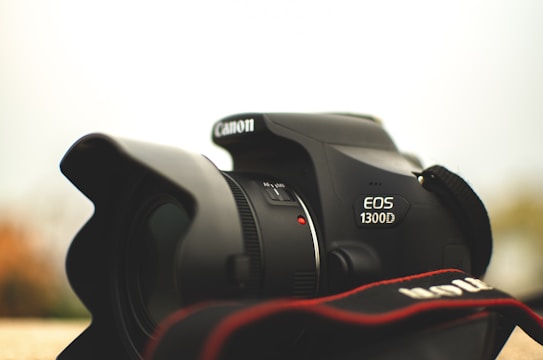 A close-up view of a Canon EOS 1300D camera, focusing on the lens and brand logo. The camera strap is partially visible in the foreground, and the background is softly blurred.