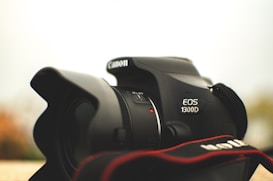 A close-up view of a Canon EOS 1300D camera, focusing on the lens and brand logo. The camera strap is partially visible in the foreground, and the background is softly blurred.