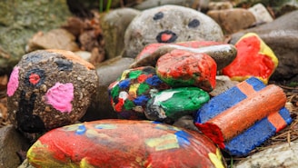 A collection of rocks painted with various bright colors and patterns is piled together. Some have faces drawn on them, while others display abstract designs. The rocks are set against a natural background of soil and stones.