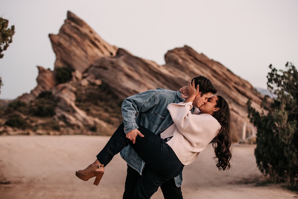 man in blue denim jacket kissing woman in white shirt on brown sand during daytime