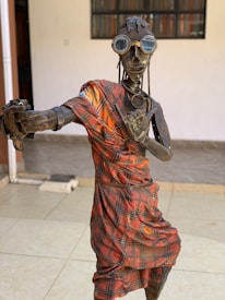 A metal sculpture of a humanoid figure wearing a plaid-patterned garment. The figure is adorned with goggles and has a distinctive, industrial look. The setting appears to be indoors with tiled flooring and a plain background.