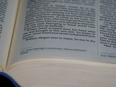 An open book features printed text on its pages, focusing on a specific passage. The text is arranged in two columns, with verse numbers marking the beginning of each passage. The edges of the pages are slightly curled, and there is some visible depth to the text block.