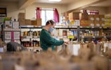 A person wearing a face mask is organizing grocery bags in what appears to be a food pantry or storage room. Shelves are filled with various boxed and canned food items. There is a window with red curtains in the background, which lets in natural light.