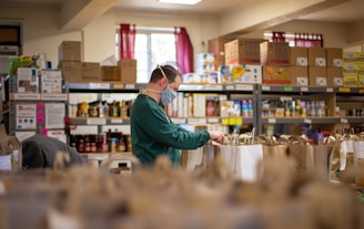 A person wearing a face mask is organizing grocery bags in what appears to be a food pantry or storage room. Shelves are filled with various boxed and canned food items. There is a window with red curtains in the background, which lets in natural light.