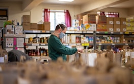 A person wearing a face mask is organizing grocery bags in what appears to be a food pantry or storage room. Shelves are filled with various boxed and canned food items. There is a window with red curtains in the background, which lets in natural light.
