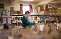 A person wearing a face mask is organizing grocery bags in what appears to be a food pantry or storage room. Shelves are filled with various boxed and canned food items. There is a window with red curtains in the background, which lets in natural light.