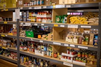 Shelves stacked with various food items including canned goods, pasta, boxed products, and bags of grains. Labels on the shelves indicate different categories of goods.