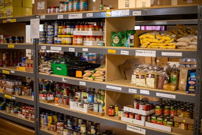 Shelves stacked with various food items including canned goods, pasta, boxed products, and bags of grains. Labels on the shelves indicate different categories of goods.