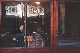 An elderly person sits behind a glass door in a room decorated with patterned textiles. A cup is placed on a table covered with a traditional rug, and natural light illuminates the scene through the glass. Outside, the reflection shows trees and part of a building.