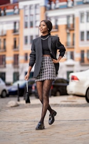 A woman confidently walks down a city street wearing a stylish ensemble including a black blazer, a patterned mini skirt, and designer patterned tights. She accessorizes with a small black handbag and black loafers, with her hair neatly styled back. The background features a blurred view of a cityscape with modern buildings and parked cars.