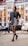 A woman confidently walks down a city street wearing a stylish ensemble including a black blazer, a patterned mini skirt, and designer patterned tights. She accessorizes with a small black handbag and black loafers, with her hair neatly styled back. The background features a blurred view of a cityscape with modern buildings and parked cars.