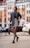A woman confidently walks down a city street wearing a stylish ensemble including a black blazer, a patterned mini skirt, and designer patterned tights. She accessorizes with a small black handbag and black loafers, with her hair neatly styled back. The background features a blurred view of a cityscape with modern buildings and parked cars.