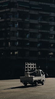 A small white truck carrying a metal frame is parked in front of a large, dark, concrete residential building. The scene is captured under low light with shadows emphasizing the structure of the building.