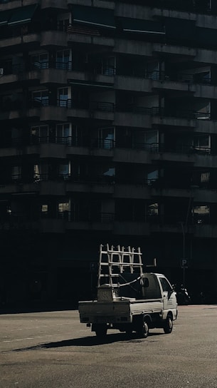 A small white truck carrying a metal frame is parked in front of a large, dark, concrete residential building. The scene is captured under low light with shadows emphasizing the structure of the building.