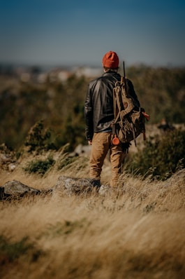 man in black jacket and brown pants wearing red knit cap standing on gray rock during