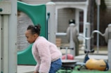 A child in a fluffy pink jacket is playing in an outdoor playground area. The child appears to be engaged with the equipment, which includes colorful structures and a play area. The background shows a residential-like setting with wooden and metal play structures.