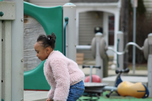 A child in a fluffy pink jacket is playing in an outdoor playground area. The child appears to be engaged with the equipment, which includes colorful structures and a play area. The background shows a residential-like setting with wooden and metal play structures.