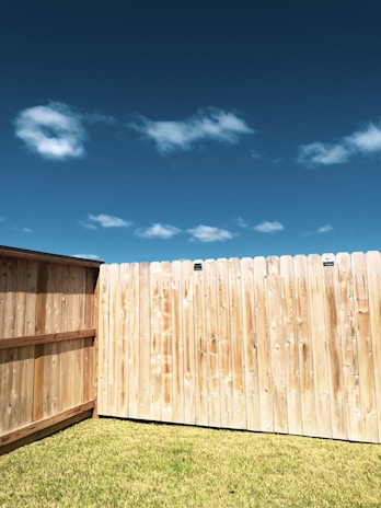 A tall wooden fence borders a grass lawn under a clear blue sky with scattered white clouds.