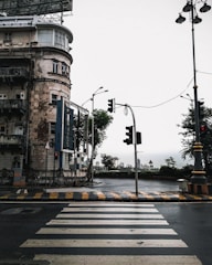 An urban scene featuring a weathered multi-story building with a large rooftop billboard structure. The street below is wet, suggesting recent rain, and features a zebra crossing and traffic lights. Street lamps and road signs are visible, with trees and city buildings visible in the background under an overcast sky.