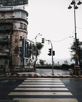 An urban scene featuring a weathered multi-story building with a large rooftop billboard structure. The street below is wet, suggesting recent rain, and features a zebra crossing and traffic lights. Street lamps and road signs are visible, with trees and city buildings visible in the background under an overcast sky.