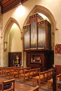 A large, ornate pipe organ is set against a stone wall, flanked by arched architectural details. In front of the organ, there are several rows of wooden chairs. To the side, a statue stands on a small table with candles and flowers.