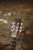 The image features the headstock of an acoustic guitar with visible tuning pegs and strings, set against a blurred earthy background. The wood of the guitar appears worn, adding a sense of vintage character.