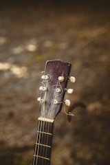 The image features the headstock of an acoustic guitar with visible tuning pegs and strings, set against a blurred earthy background. The wood of the guitar appears worn, adding a sense of vintage character.