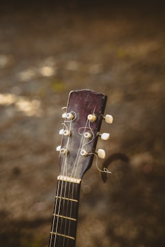 The image features the headstock of an acoustic guitar with visible tuning pegs and strings, set against a blurred earthy background. The wood of the guitar appears worn, adding a sense of vintage character.