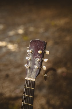 The image features the headstock of an acoustic guitar with visible tuning pegs and strings, set against a blurred earthy background. The wood of the guitar appears worn, adding a sense of vintage character.