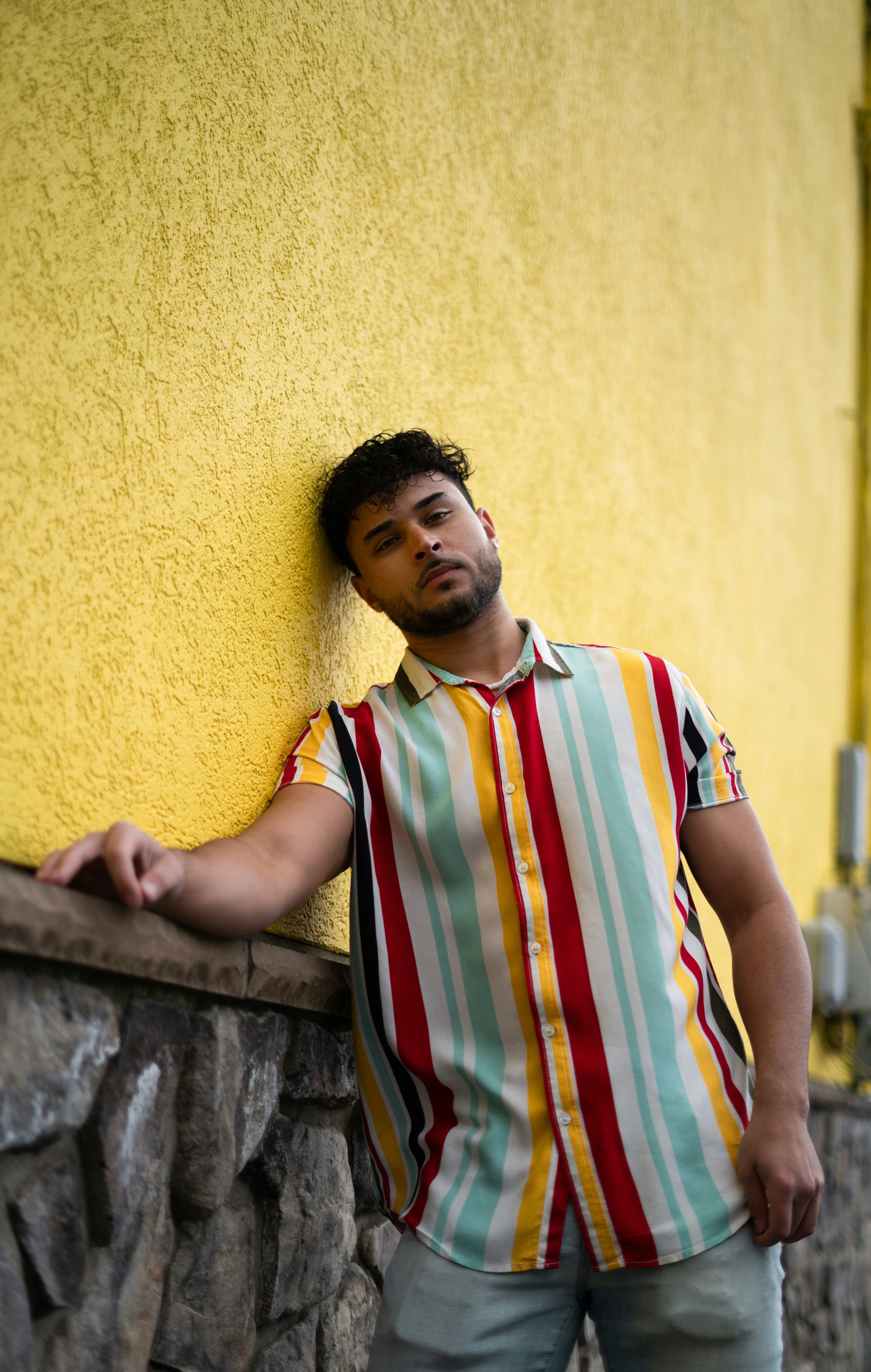 man in white red and blue stripe polo shirt leaning on yellow concrete wall during daytime