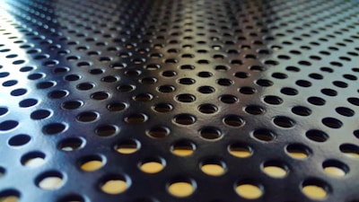 A close-up of a metallic surface with a repeating pattern of circular holes. The pattern creates a sense of depth and texture, with light reflections visible on the shiny surfaces.