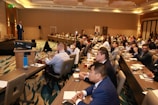 A conference room filled with people sitting at tables, attentively listening to a speaker on stage. The room is well-lit with modern decor, and attendees have notebooks and drinks on the tables.