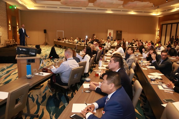 A conference room filled with people sitting at tables, attentively listening to a speaker on stage. The room is well-lit with modern decor, and attendees have notebooks and drinks on the tables.