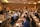 A conference room filled with people sitting at tables, attentively listening to a speaker on stage. The room is well-lit with modern decor, and attendees have notebooks and drinks on the tables.