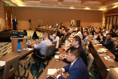 A conference room filled with people sitting at tables, attentively listening to a speaker on stage. The room is well-lit with modern decor, and attendees have notebooks and drinks on the tables.