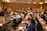 A conference room filled with people sitting at tables, attentively listening to a speaker on stage. The room is well-lit with modern decor, and attendees have notebooks and drinks on the tables.