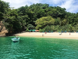 A serene beach scene with turquoise waters and a small boat floating in the foreground. The sandy shore is lined with a few people walking and a cluster of umbrellas and chairs. Behind the beach, lush green trees form a dense forest, creating a tranquil, secluded atmosphere. The sky is partly cloudy, adding layers to the tranquil landscape.