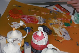 A collection of paint bottles with caps open and various colors smeared, mixed on an artist's palette. Behind, there are colored pencils or markers in a box and a hand holding a piece of paper or canvas. The setting suggests an art studio or workspace.