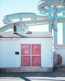 A small white building with a faded red door is positioned in front of a series of large water slides. The building has a chimney and a small vent on its roof. A chain-link fence is partially visible to the right. The water slides are light blue and twist in the background under a clear blue sky.
