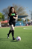 woman in black and green nike soccer jersey kicking soccer ball on green grass field during