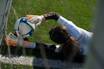 A soccer goalkeeper in a white long-sleeve shirt and gloves is seen diving on a green grass field, stretching to catch a soccer ball. The scene is viewed from an elevated angle, near a goalpost, with the player's hair visible on the ground.