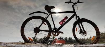 A bicycle is silhouetted against a sky with a sun partially obscured by clouds. There is a water bottle attached to the frame of the bicycle. In the background, a row of palm trees and a vehicle can be seen.