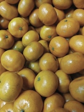 A cluster of fresh, ripe oranges fills the entire image. The oranges are closely packed, showcasing their smooth, shiny skin and vibrant orange color. A few have small green spots at the top where the stem is attached.