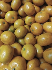 A cluster of fresh, ripe oranges fills the entire image. The oranges are closely packed, showcasing their smooth, shiny skin and vibrant orange color. A few have small green spots at the top where the stem is attached.