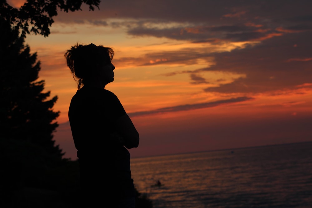 silhouette of woman standing near body of water during sunset