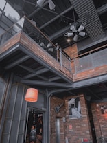 A modern industrial-style interior with exposed brick walls and metal beams. There is a hanging light fixture and a staircase leading to an upper level with a metal railing. Various photographs and a guitar-shaped decoration are mounted on the walls.