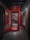 A vibrant red phone booth stands open in a dimly lit corridor with brick walls. Inside the booth, a mural featuring a face with colorful abstract designs is visible. The area is industrial with exposed beams and a single hanging light fixture to the right.
