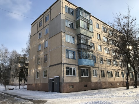 A five-story apartment building with a simple, rectangular design is set against a winter backdrop. The building features brickwork with horizontal lines dividing the floors, and balconies with glass and blue panels. Snow covers the ground and bare trees stand nearby, with sunlight filtering through the branches.