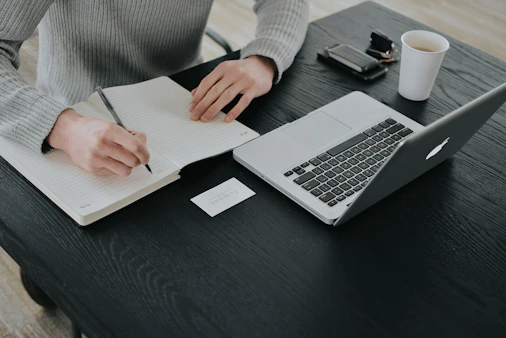 A person in a grey sweater writing in a notebook at a black desk