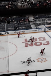 A hockey game scene featuring players in red and black jerseys skating on an ice rink. The rink is set inside an arena with partially occupied spectator stands. Visible advertisements along the rink boards indicate sponsorships.